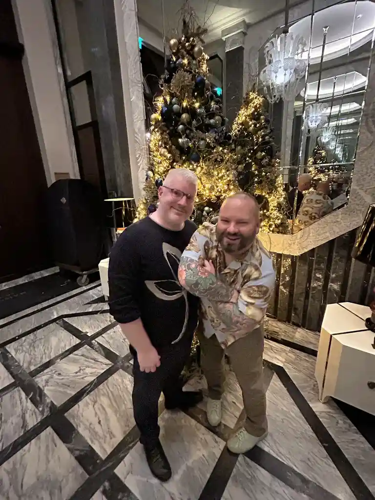 Two men pose for a photo in a luxurious lobby with a large, ornate Christmas tree decorated with gold and blue ornaments and…