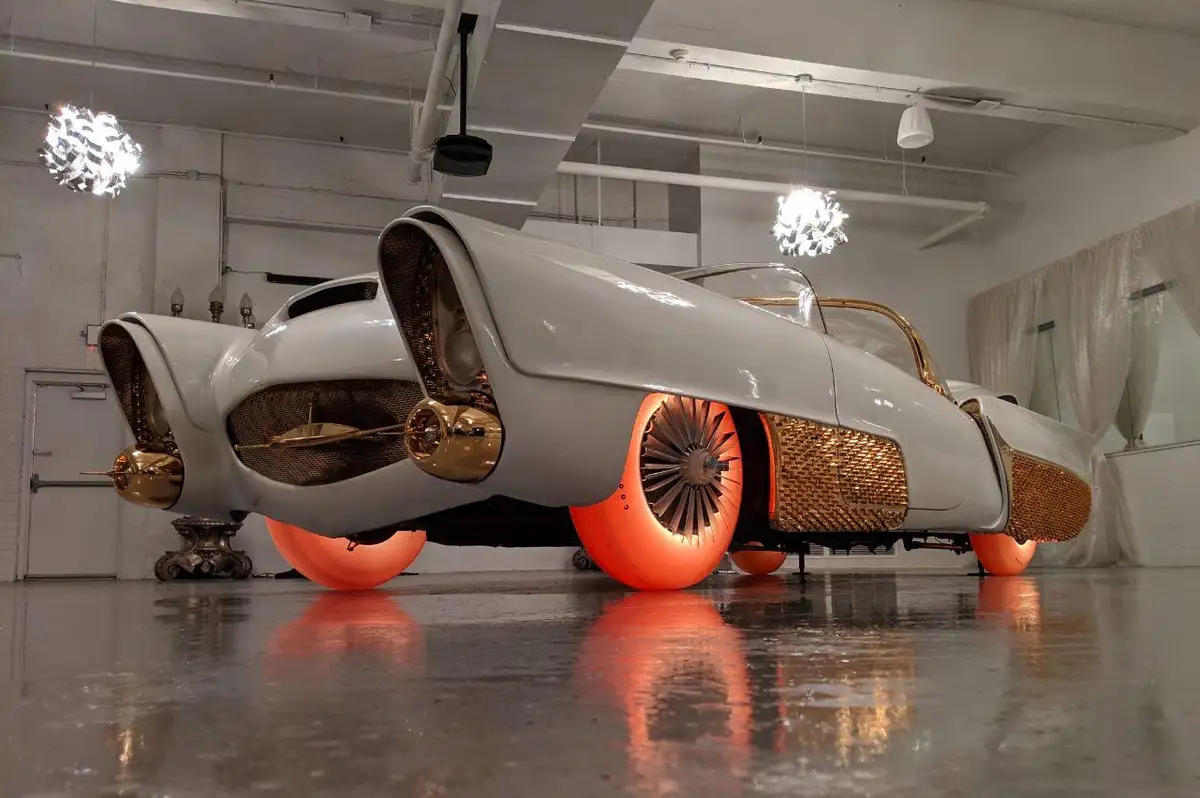 a low-angle shot of a white, futuristic-looking car with large orange wheels and gold trim, displayed indoors on a…