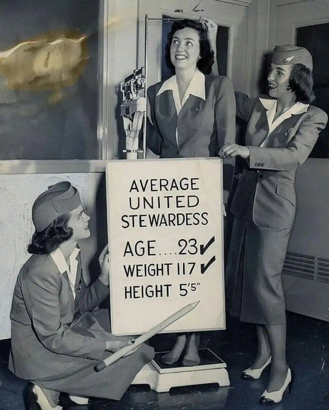 A vintage black-and-white photo shows three women in 1940s-style airline stewardess uniforms. One woman stands on a scale…