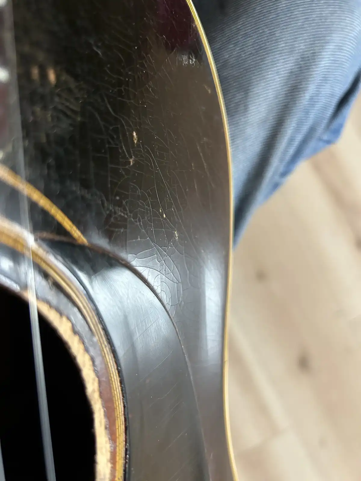 a close-up photo shows the worn, cracked finish of a dark-colored acoustic guitar's body, with gold trim and a visible…