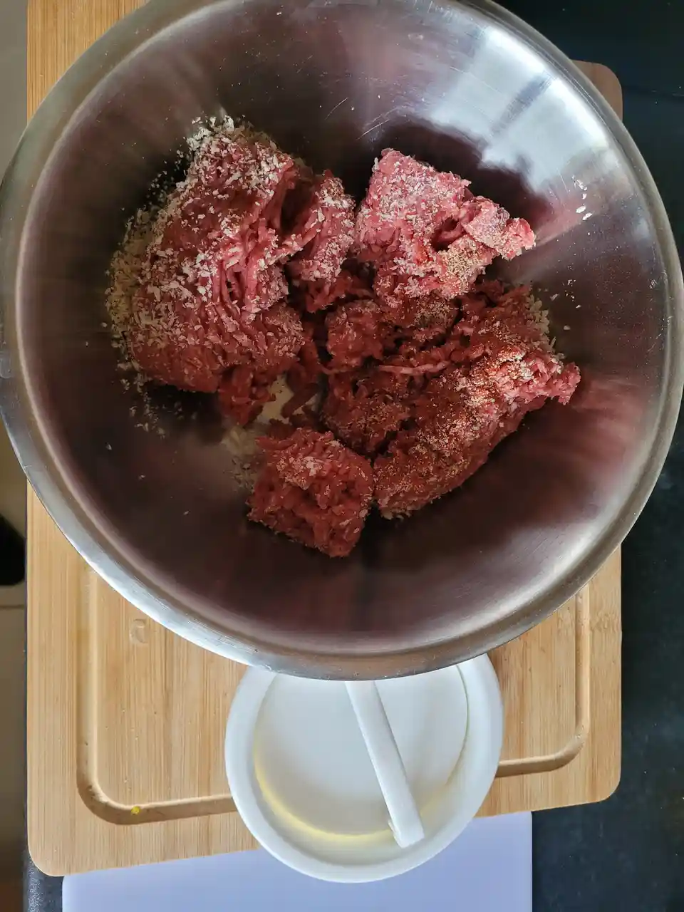 A stainless steel mixing bowl filled with raw ground meat and breadcrumbs sits atop a white plastic cup with a straw, placed…
