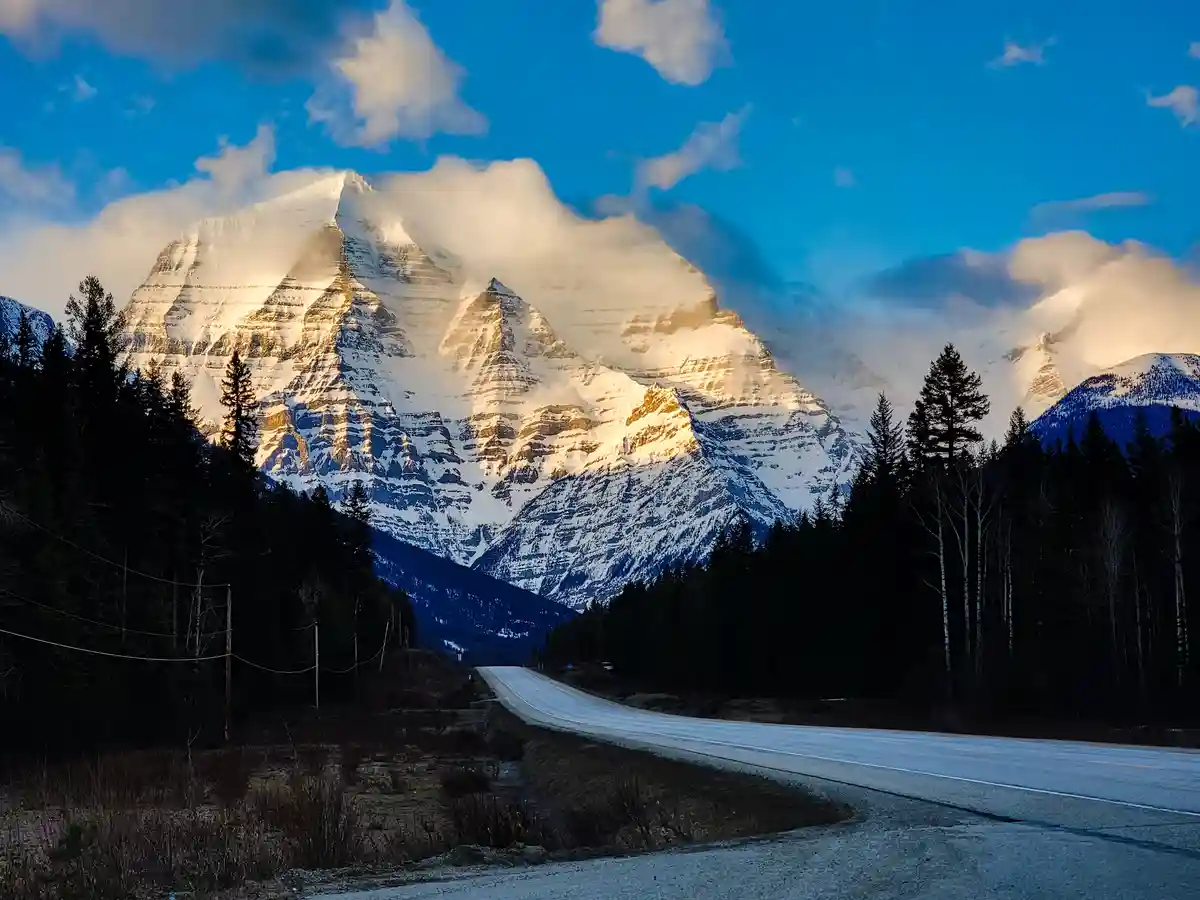 A scenic photo shows a winding road leading toward majestic, snow-capped mountains under a blue sky with clouds. The…