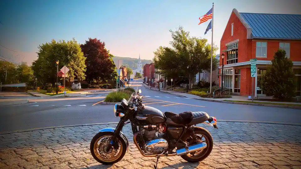 A black Triumph Bonneville T120 motorcycle is parked on a cobblestone street in front of a red brick building labeled…