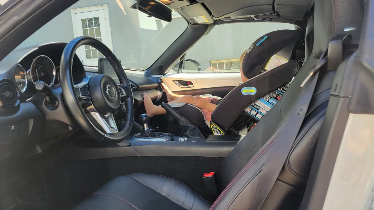 A child is seated in a car seat inside a Mazda sports car, with their bare feet resting on the center console near the gear…