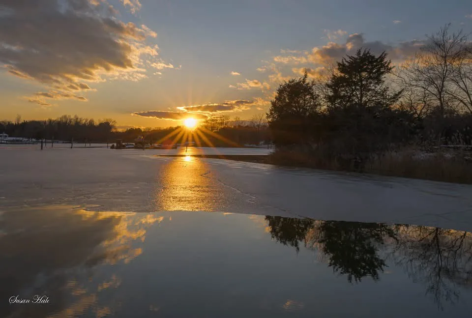 A serene winter sunset over a partially frozen lake, with the sun low on the horizon casting golden rays and reflecting in…