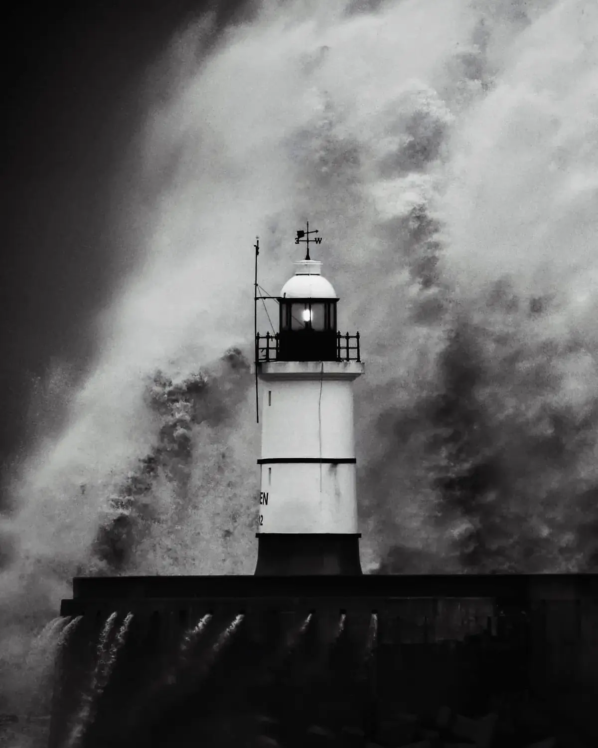 A black-and-white photo shows a lighthouse standing firm as a massive wave crashes around it, spraying water high into the…