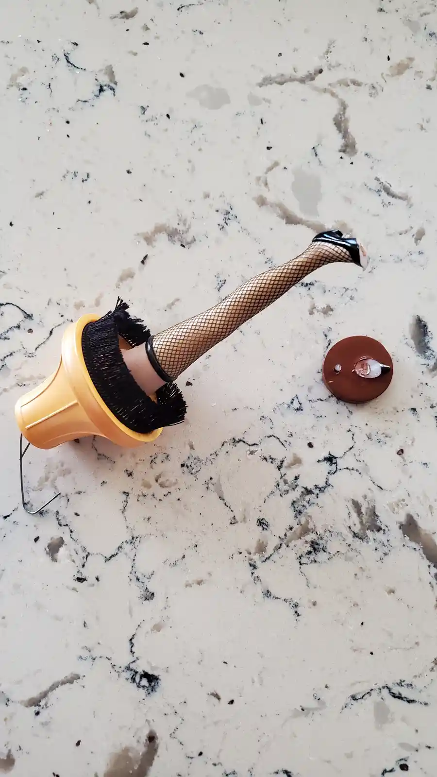 A yellow toilet brush with a black bristle head lies on a speckled countertop, its handle replaced by a doll's leg wearing…