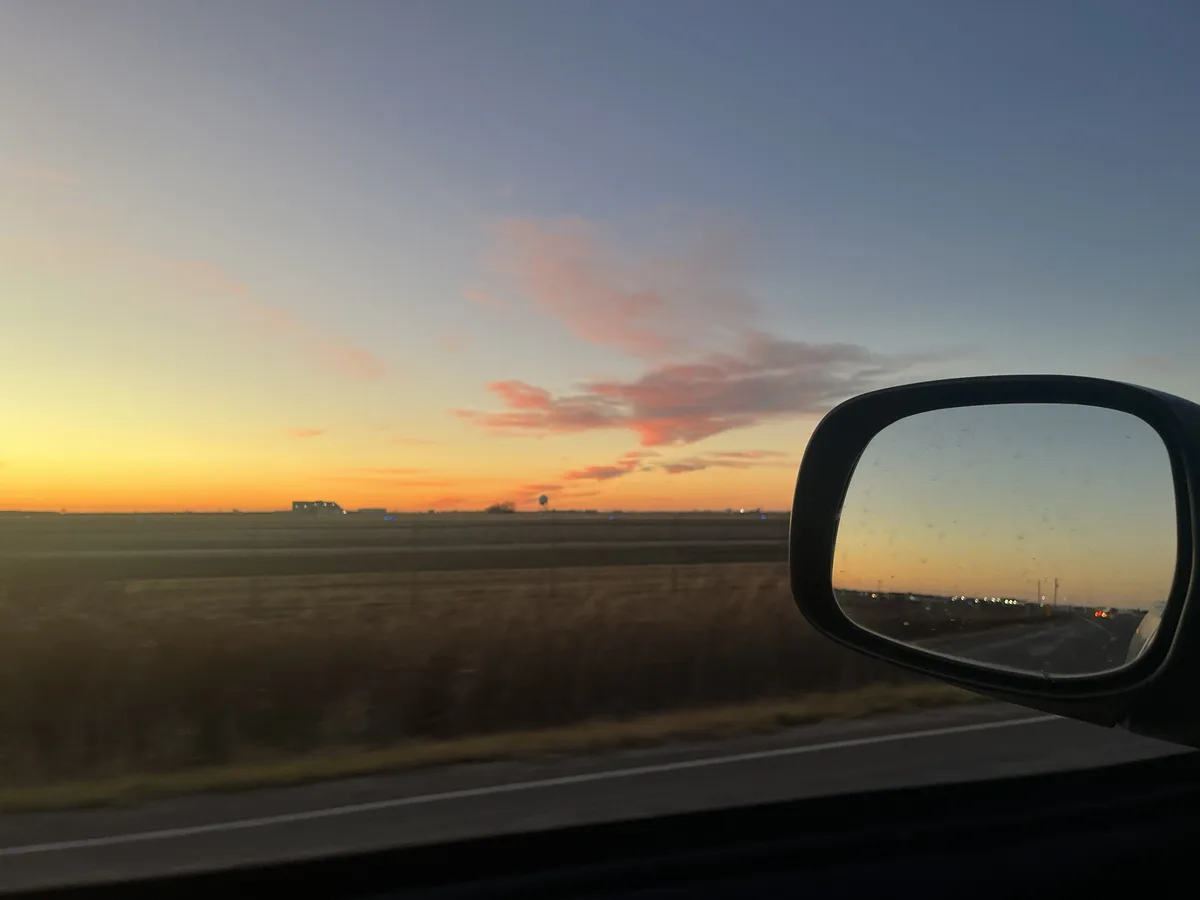 A photo taken from inside a moving car shows a beautiful sunset with orange and pink clouds over a flat landscape. The car's…