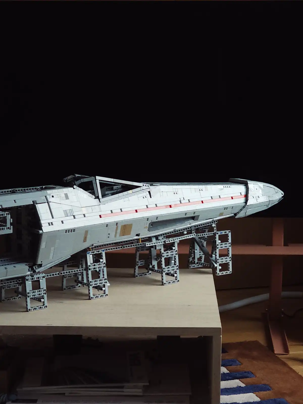 A detailed Lego model of a spaceship, resembling a Star Wars X-wing fighter, is displayed on a custom-built Lego stand on a…