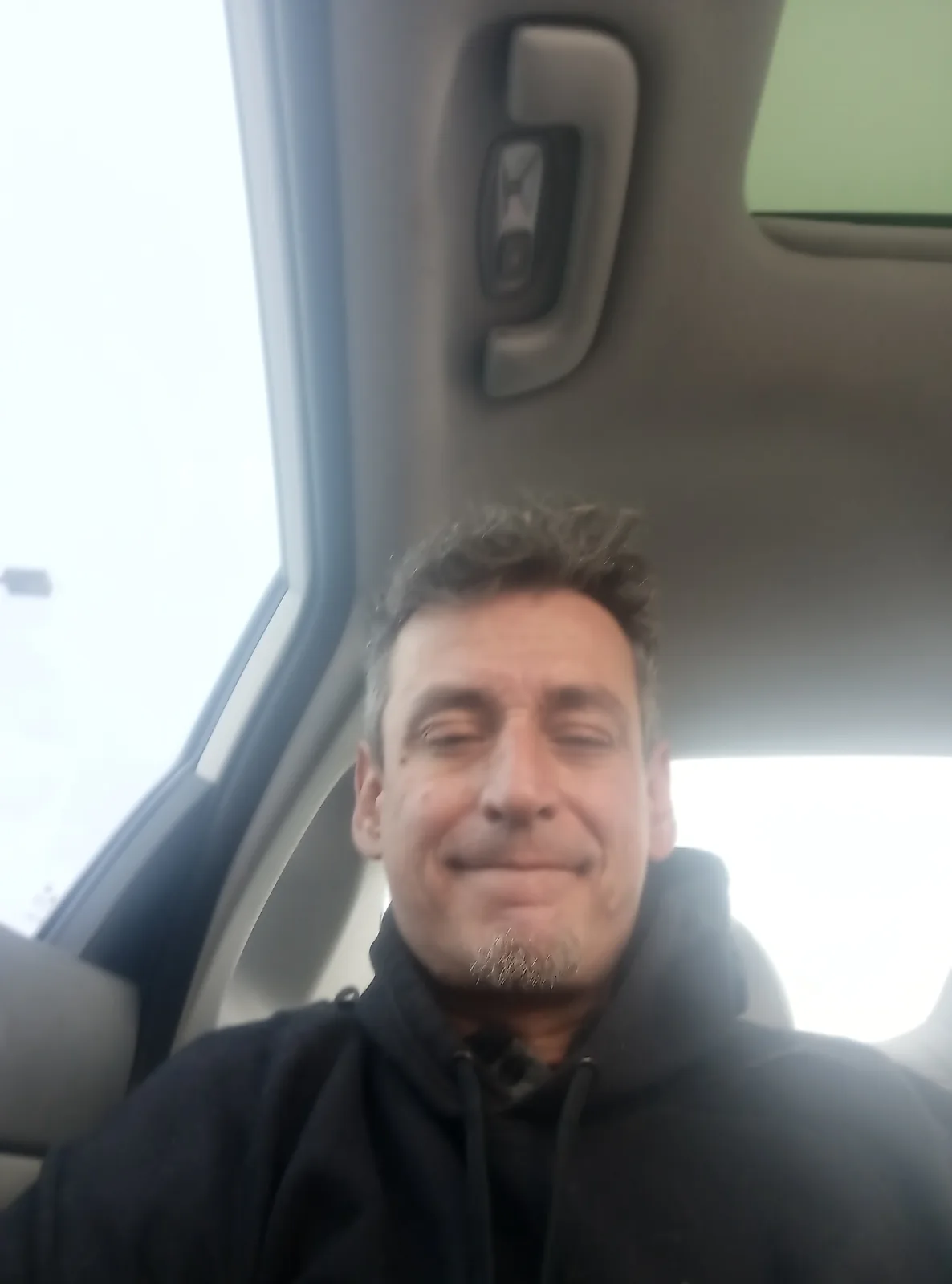 A man with short dark hair and a goatee, wearing a black hoodie, sits inside a car and smiles with his eyes closed. The…