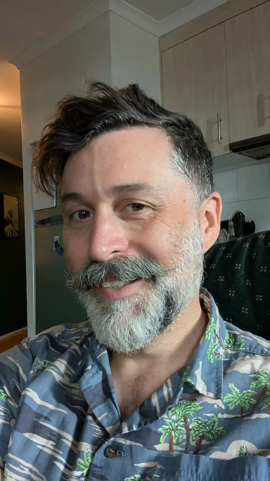 A middle-aged man with a salt-and-pepper beard and mustache smiles at the camera while wearing a blue Hawaiian shirt with…