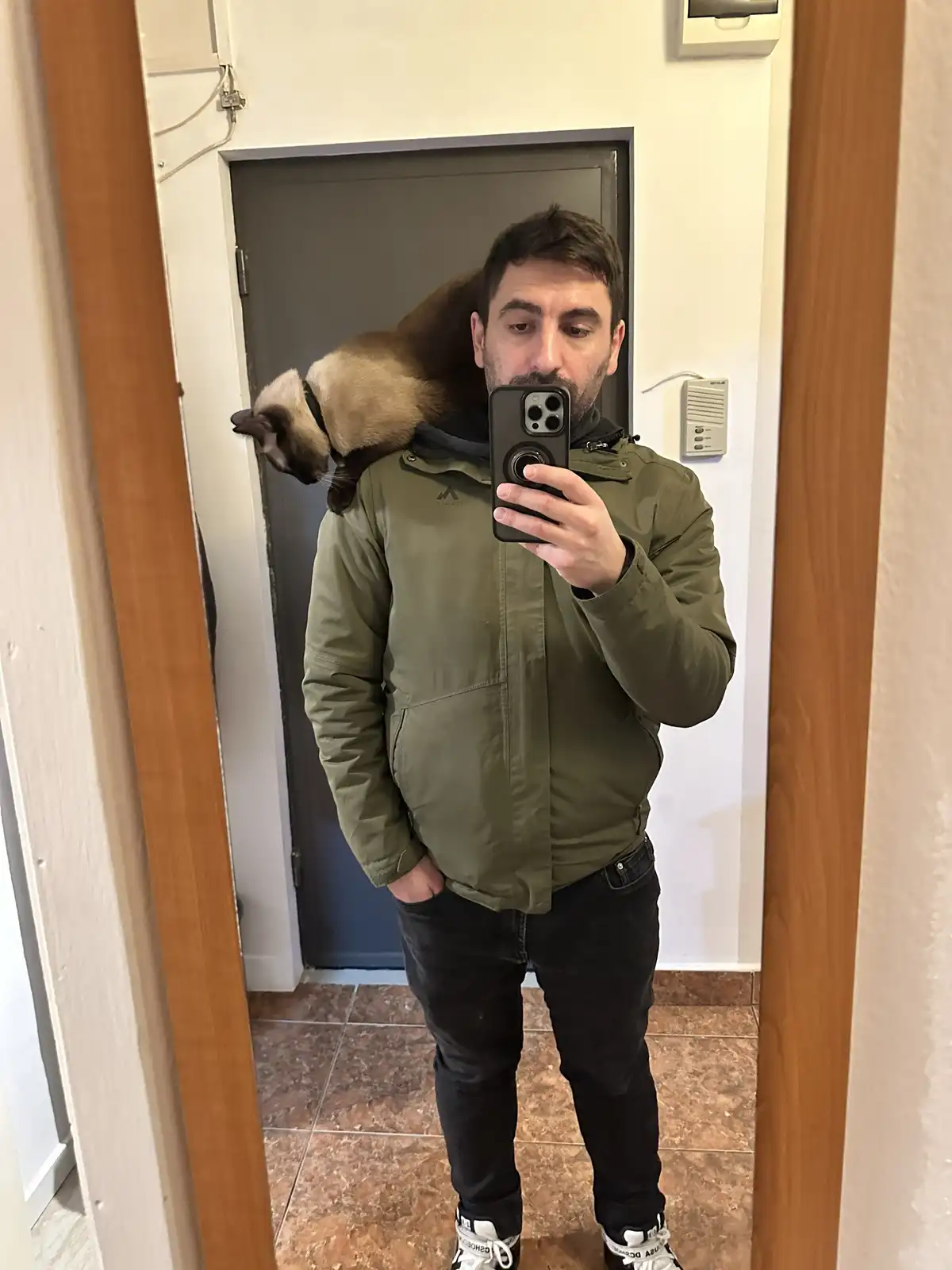 A man in an olive green jacket takes a mirror selfie while a Siamese cat rests on his shoulders, looking toward the camera…