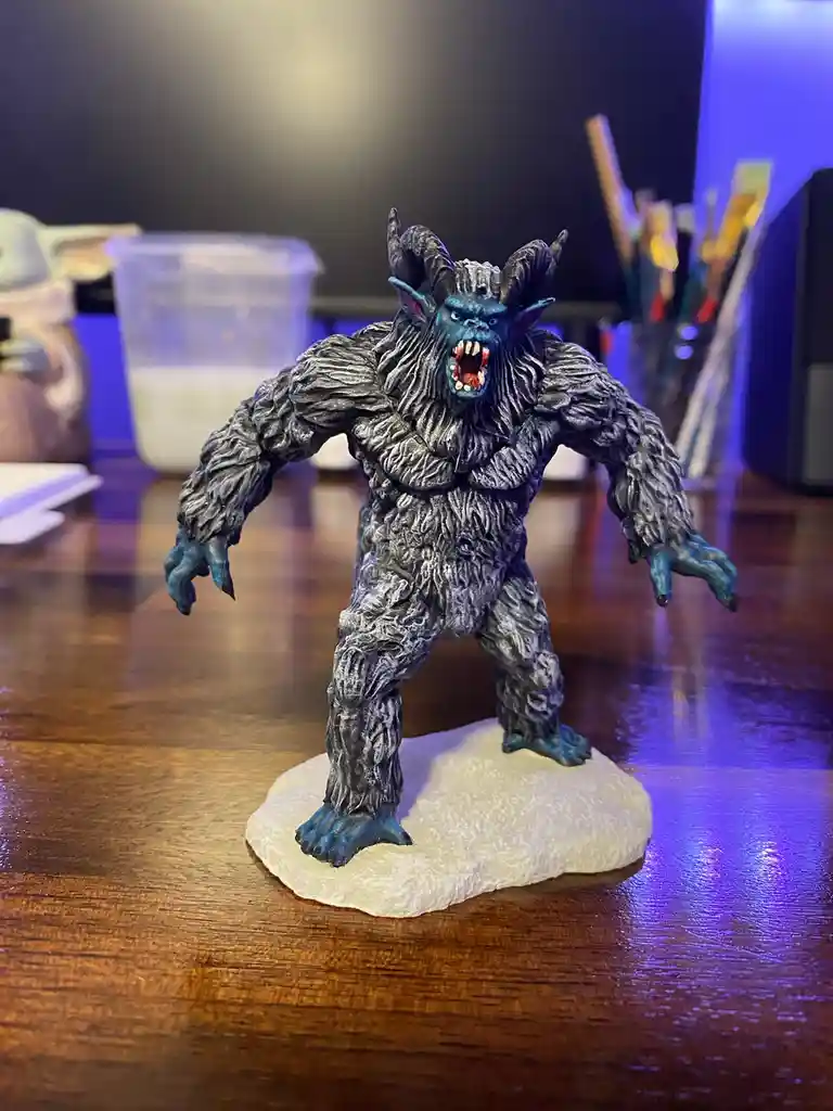 A painted miniature figurine of a monstrous, blue-skinned creature with horns, sharp teeth, and fur, standing on a white…