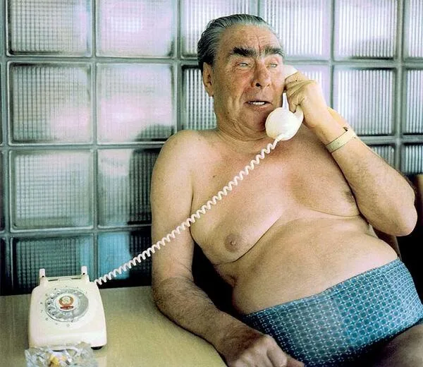 A shirtless, overweight man in blue patterned underwear sits at a table holding a white rotary phone to his ear, looking…