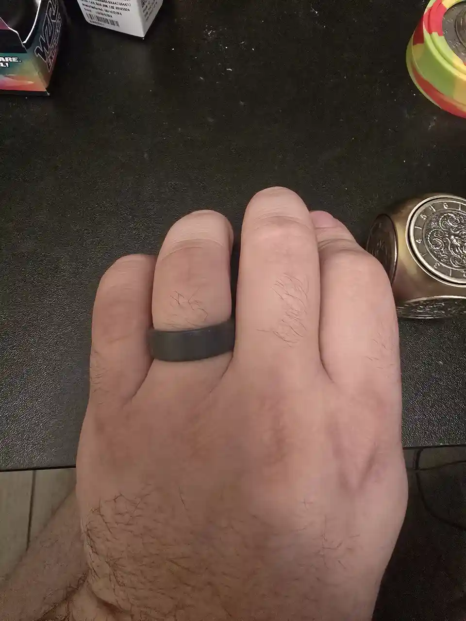 A close-up photo shows a person's hand resting on a dark surface, with a simple black ring on the ring finger. The hand has…