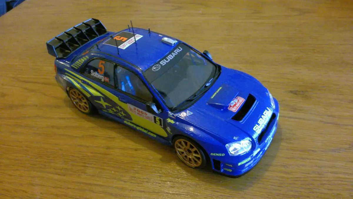 A blue Subaru rally car model with yellow decals and the number 5, sitting on a wooden table. The car has 'Solberg' and…