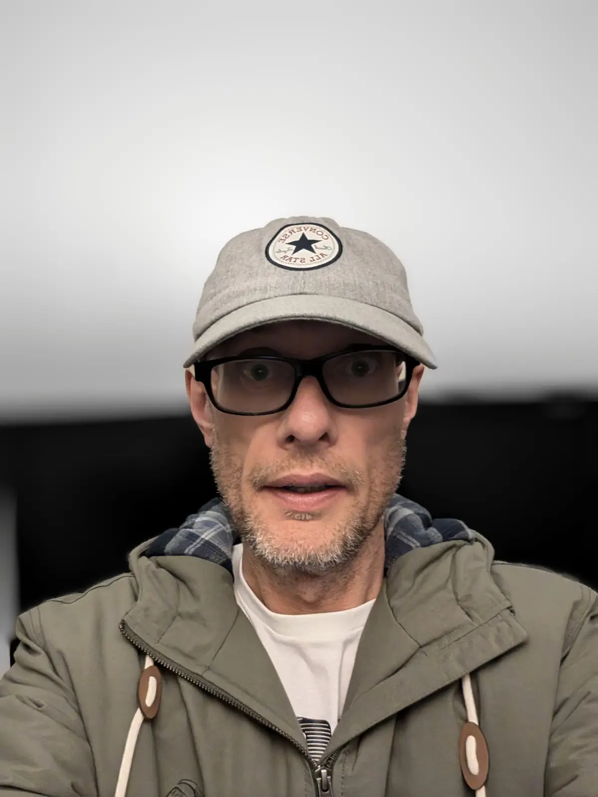 A man with a gray Converse All Star cap, black-rimmed glasses, and a surprised expression stares directly at the camera. He…