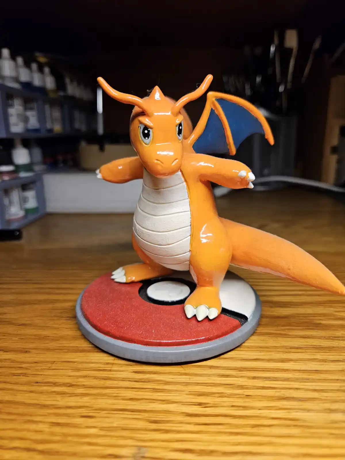 A handmade orange dragon figurine, resembling Charizard from Pokémon, stands on a red and white Poké Ball base with its arms…