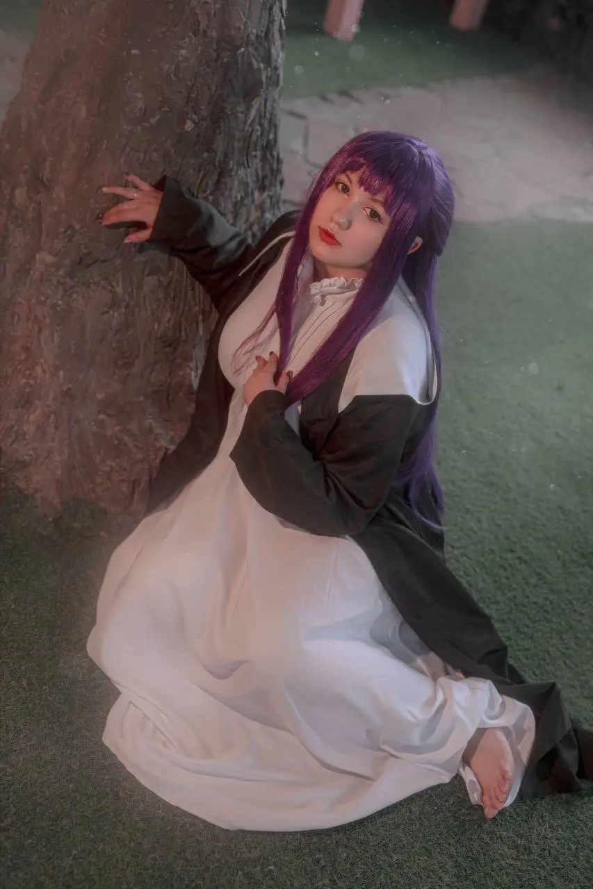 A person with long purple hair and red lipstick is kneeling on grass next to a tree, wearing a white dress with a black…