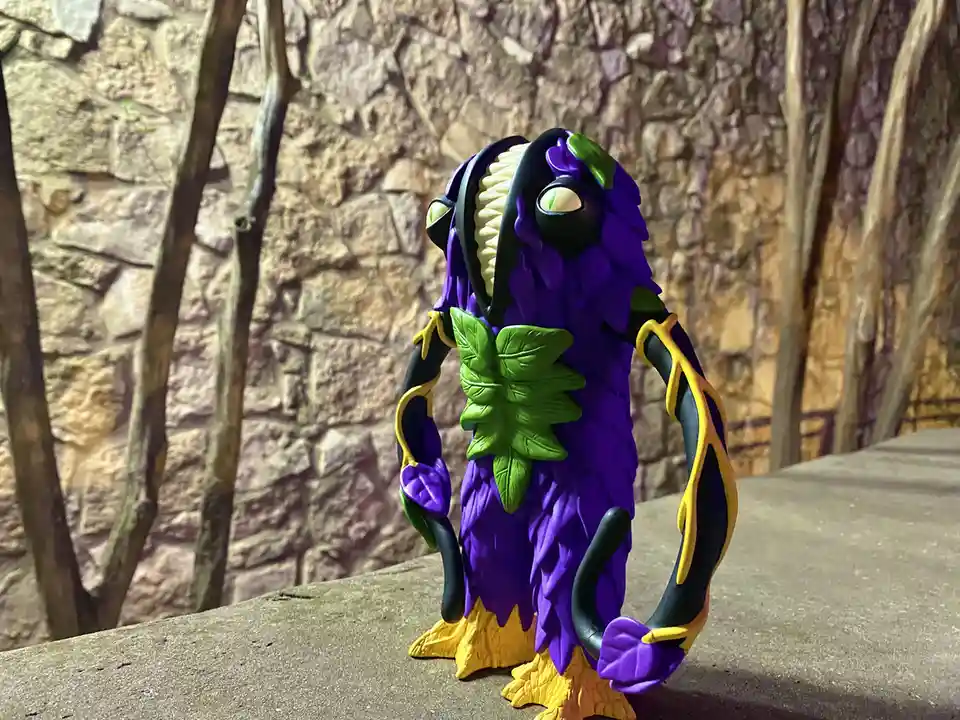 A purple, plant-like monster toy with green leaves and yellow accents stands on a concrete ledge against a stone wall and…