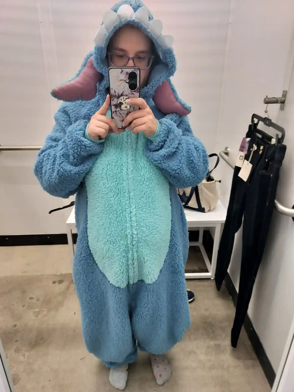 A person wearing a fluffy blue and teal Stitch-like onesie with purple ears and white teeth on the hood, takes a mirror…