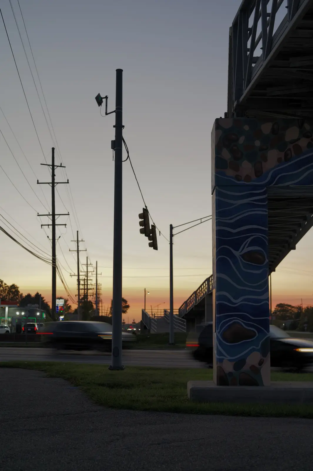 A photograph of a street scene at dusk with a traffic light pole and a painted bridge pillar. Cars are blurred in motion on…