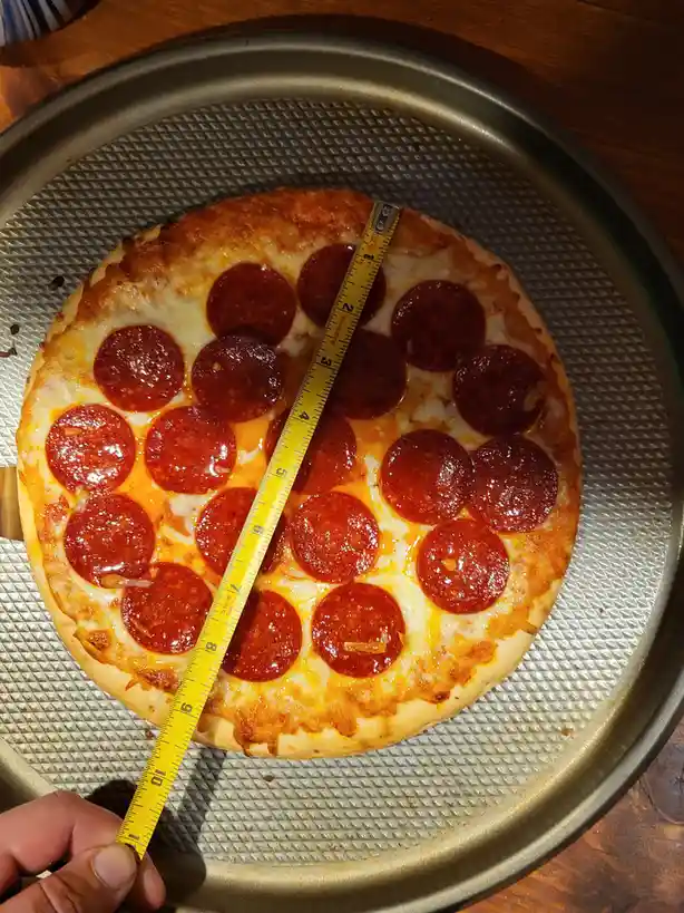A freshly baked pepperoni pizza sits on a metal baking tray, with a yellow measuring tape stretched across its diameter…