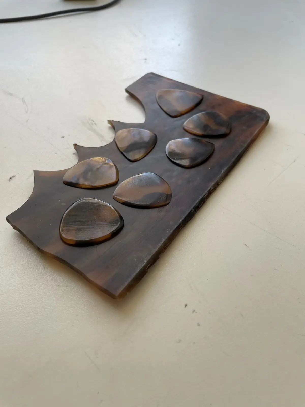 A close-up photo shows a dark brown, tortoiseshell-patterned guitar pick holder shaped like a guitar pick, with eight actual…