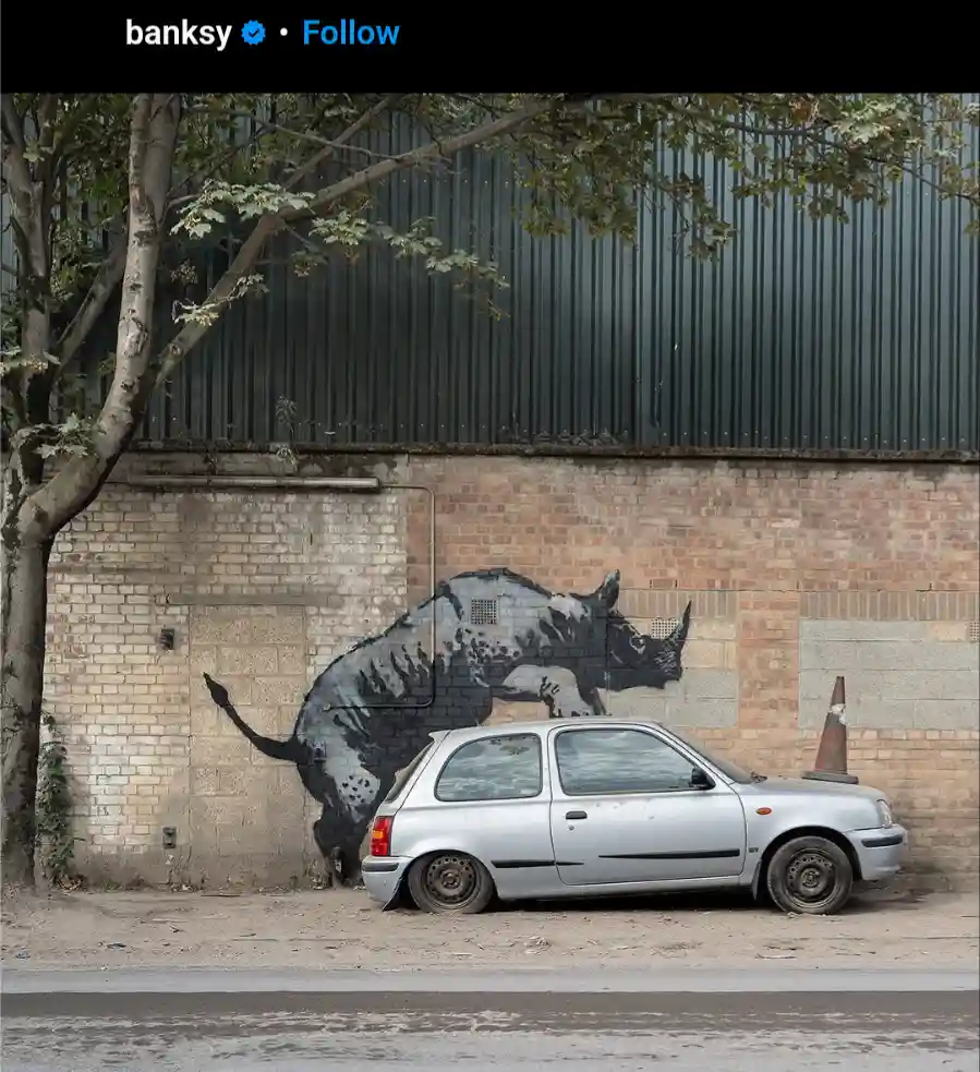 A silver, dilapidated car is parked in front of a brick wall with a large Banksy mural of a rhinoceros seemingly charging…