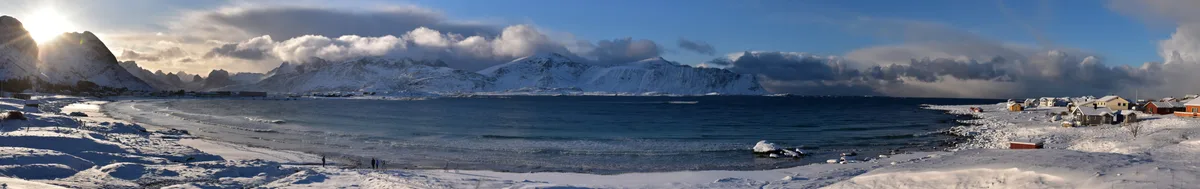 A wide, snowy coastal panorama shows a frozen bay with snow-covered mountains in the background and colorful houses along…