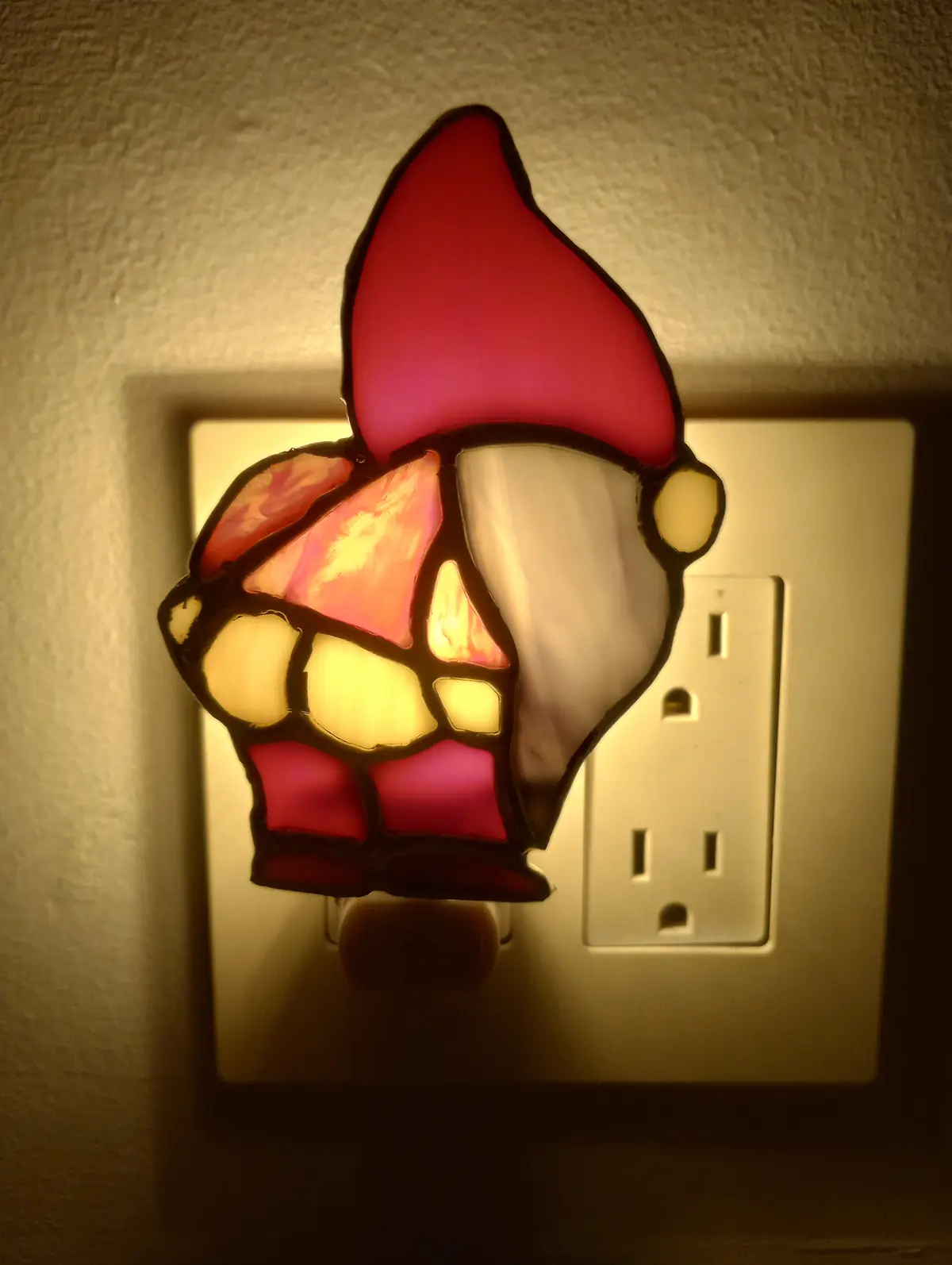 A stained-glass nightlight shaped like a gnome with a red hat and white beard is plugged into a wall outlet, glowing warmly…
