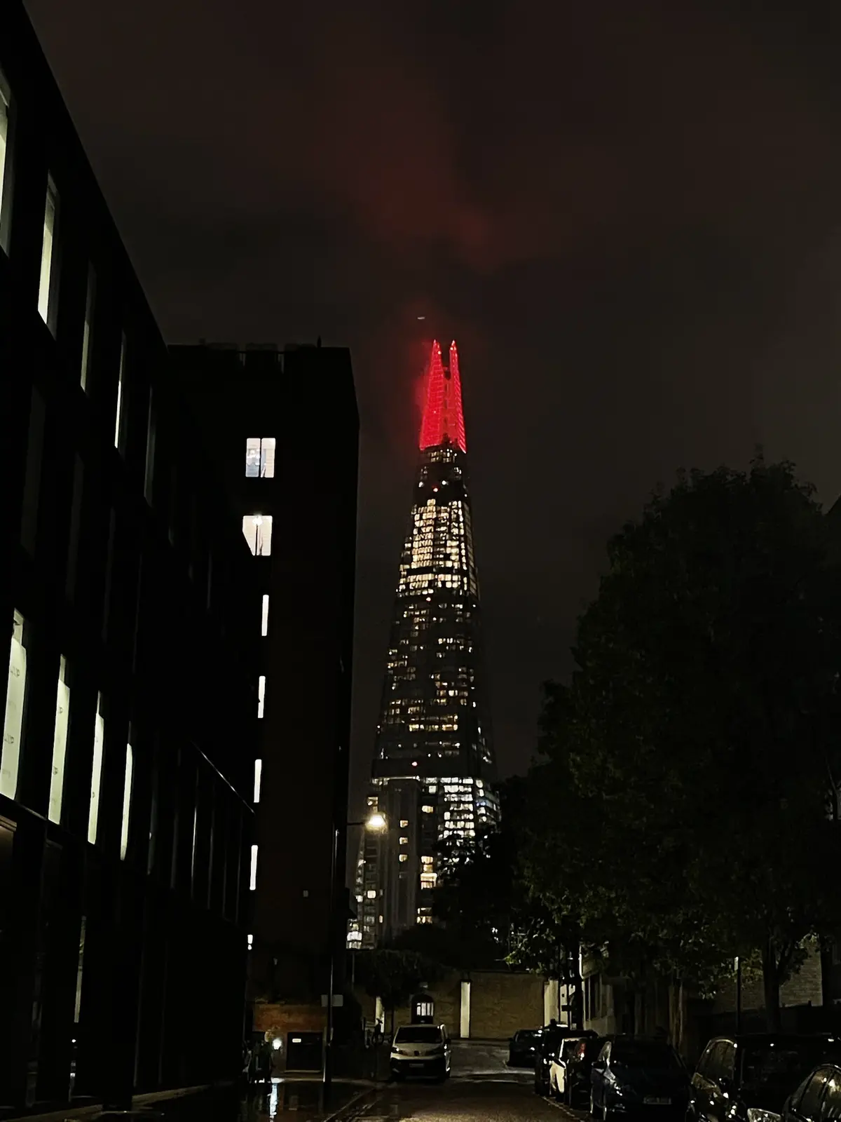 A nighttime street view looking up at the Shard skyscraper in London, which is illuminated with red lights at its spire…