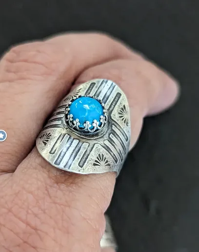 A close-up photo shows a hand wearing a large, ornate silver ring with a bright blue turquoise stone set in the center. The…