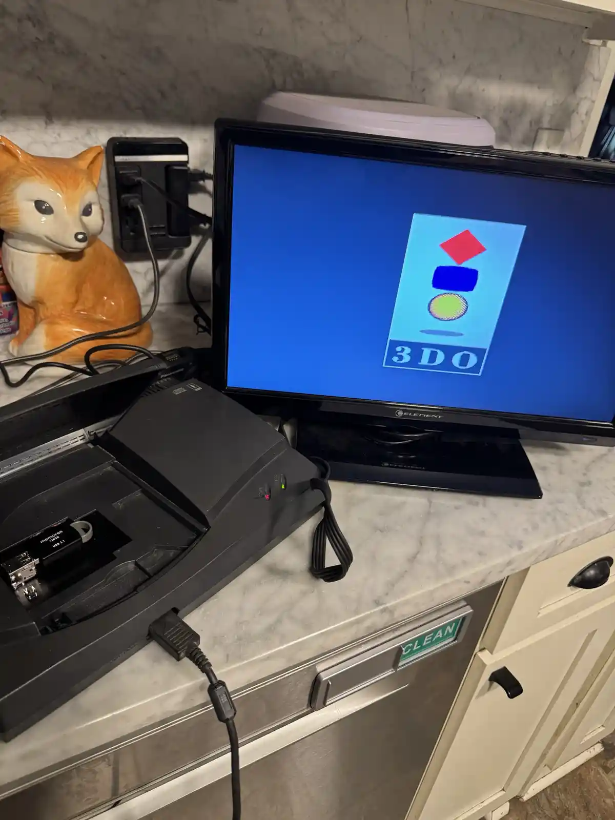 A black 3DO gaming console sits on a marble countertop, its lid open to reveal a USB drive inside, connected to a small TV…