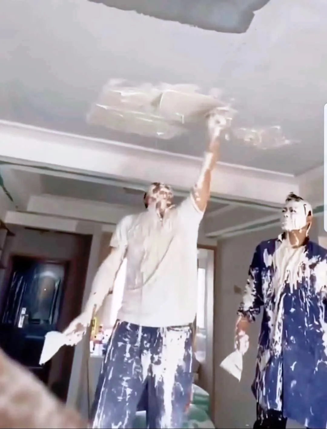 Two men covered in white plaster are working on a ceiling; one is applying plaster with a trowel while the other stands…
