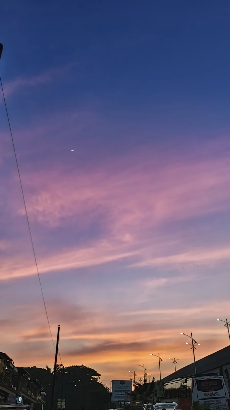 A photo of a twilight sky with a crescent moon and pink-purple clouds above a street with buildings, trees, and…