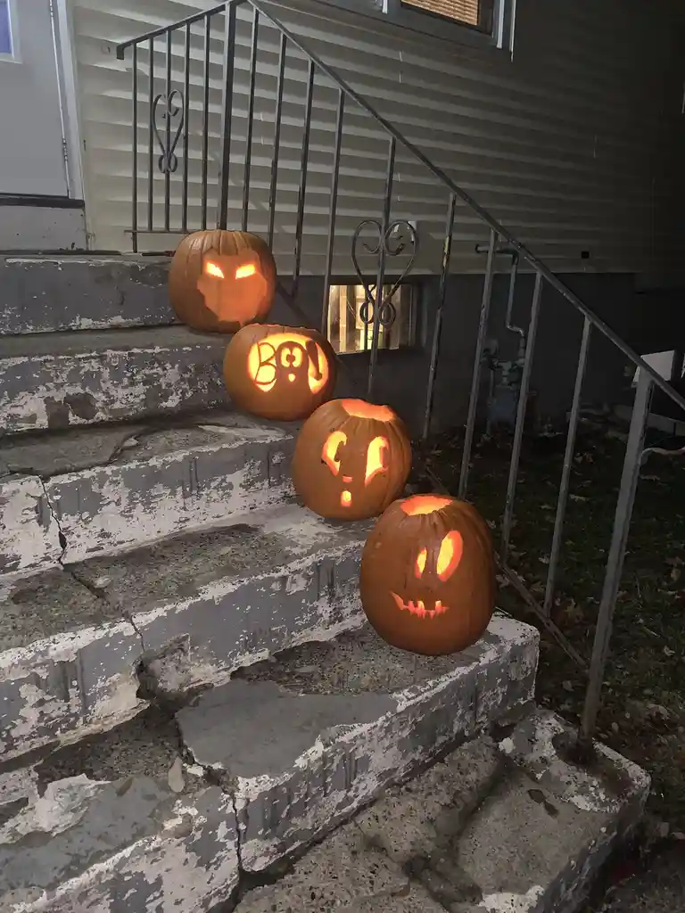 Four carved pumpkins with glowing faces sit on weathered concrete steps leading to a house, with one pumpkin in the middle…