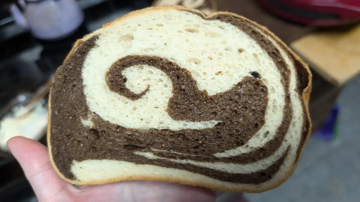 A close-up photo shows a hand holding a slice of swirl bread with alternating light and dark brown dough, forming a spiral…