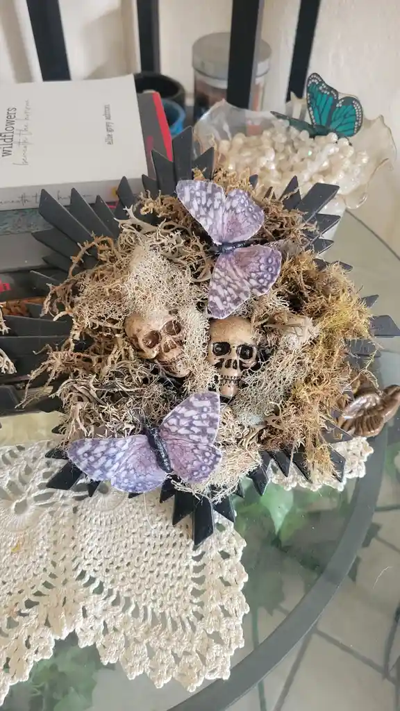 A decorative arrangement resembling a hedgehog made of black plastic spikes, dried moss, and two small skulls, adorned with…