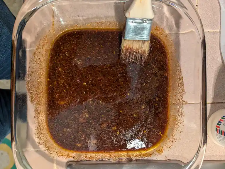 A top-down photo shows a paintbrush dipped in a dark, speckled liquid inside a clear glass baking dish, with the brush…