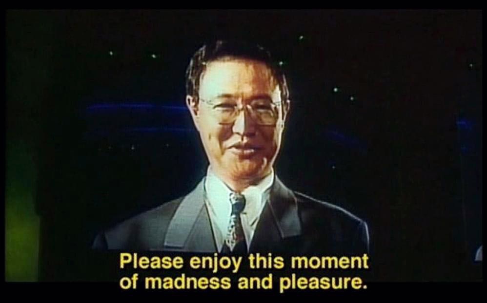A still image of a man in a suit and glasses, smiling slightly against a dark background with faint blue lights. Yellow text…
