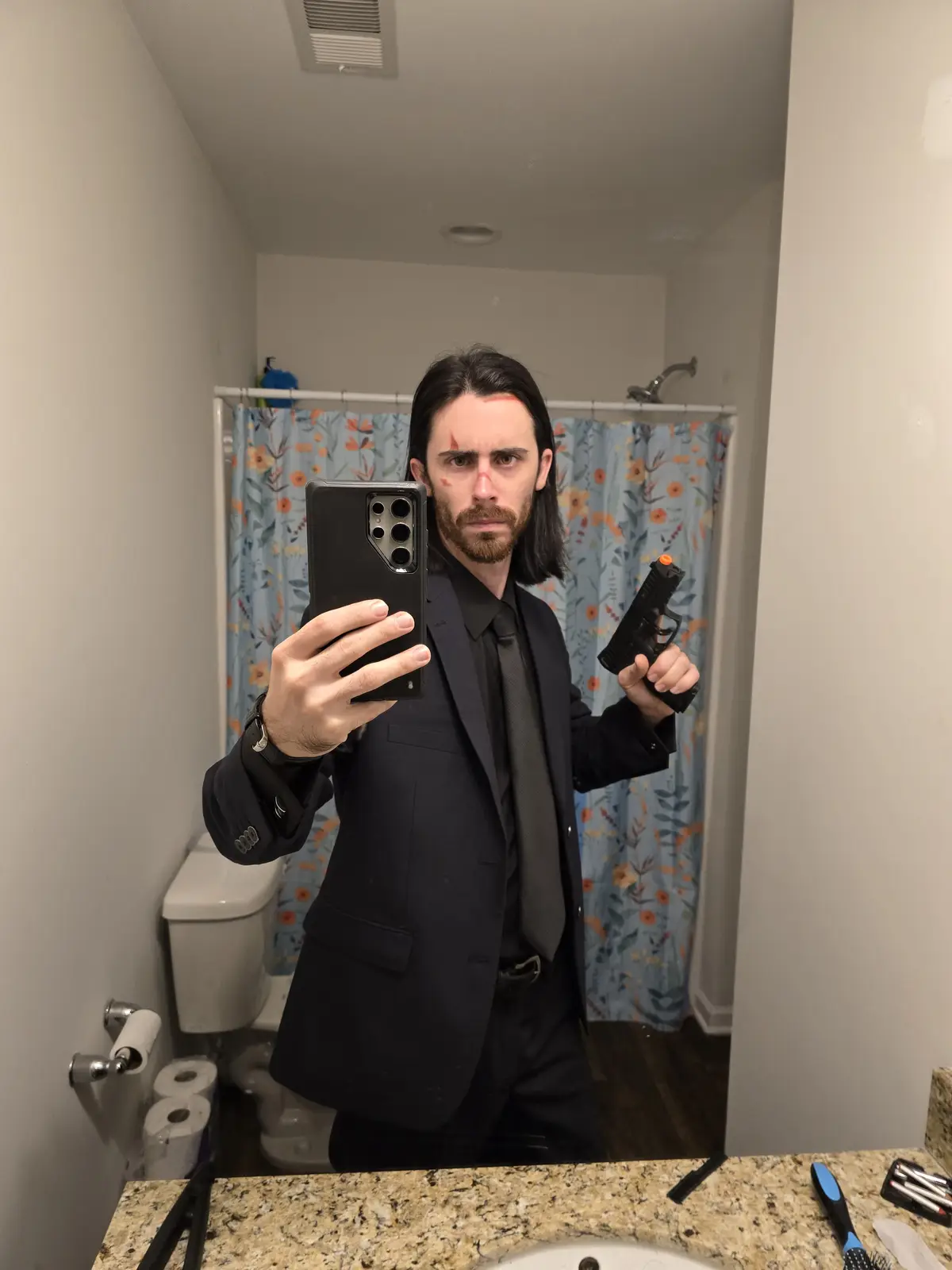 A man in a black suit and tie with fake blood on his face takes a mirror selfie in a bathroom while holding a toy gun. He…