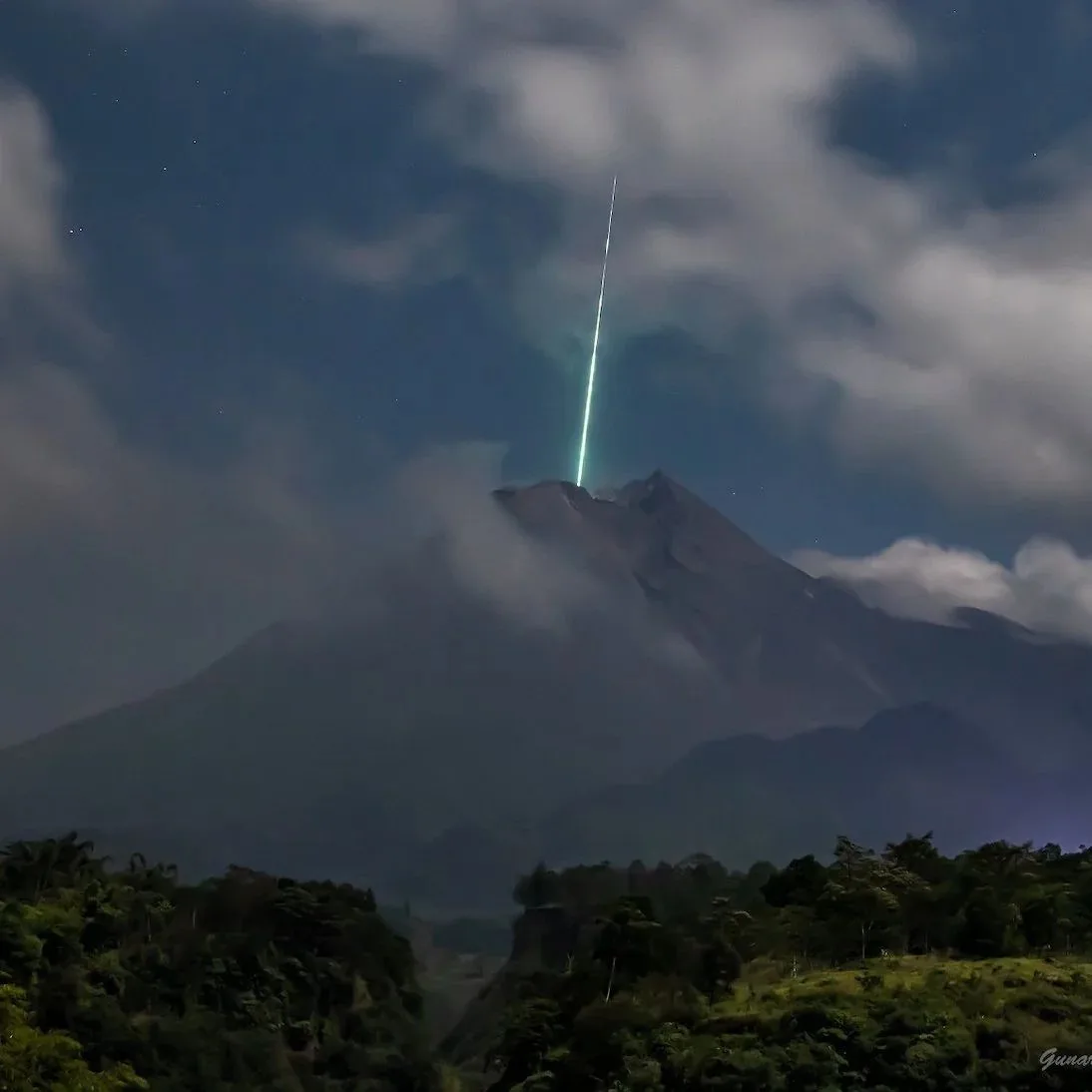 A night photograph shows a large, dark mountain peak partially obscured by clouds, with a bright, vertical green laser beam…