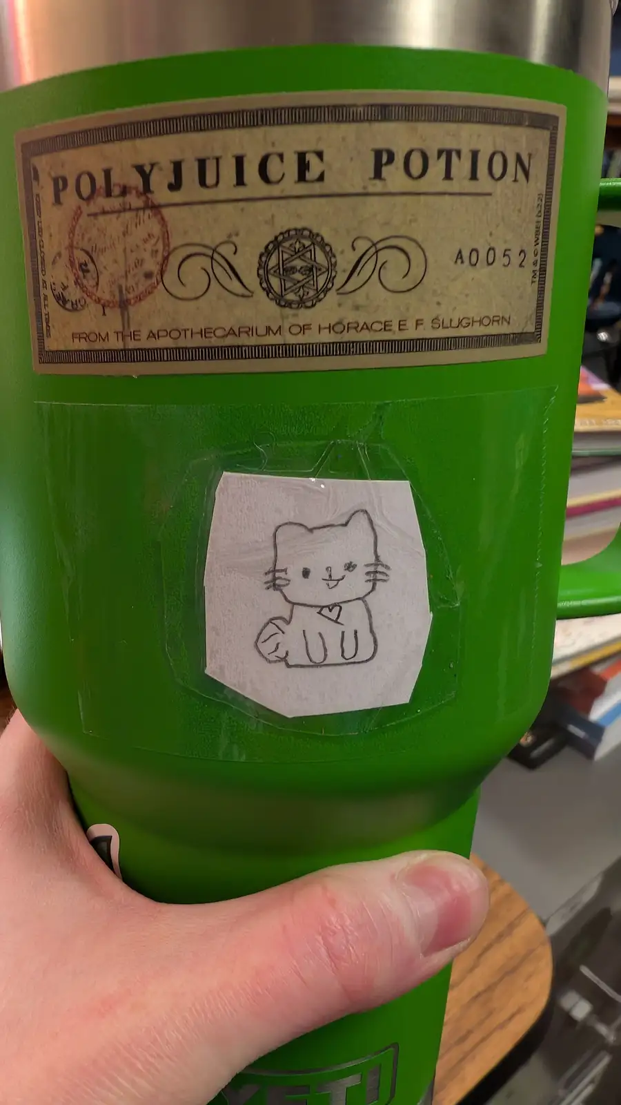 A green Yeti tumbler is labeled as a 'Polyjuice Potion' from Horace Slughorn's apothecarium, with a hand-drawn cartoon cat…