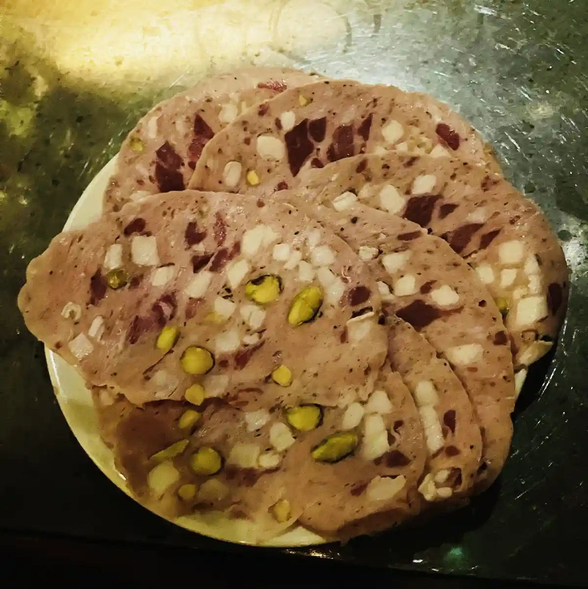 A close-up photo shows several slices of terrine arranged on a white plate, resting on a dark, textured surface. The terrine…