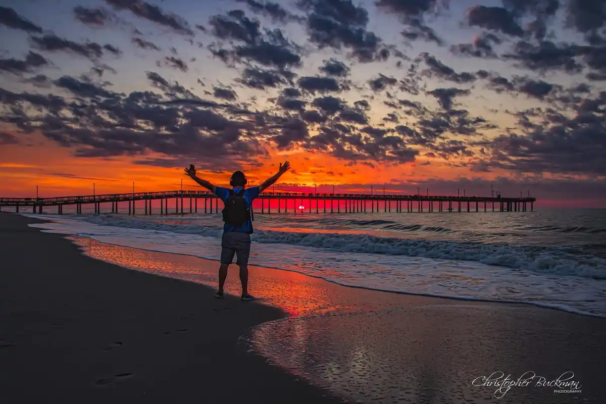 A person stands on a beach at sunset with arms raised in joy, wearing headphones and holding a phone, facing a pier and…