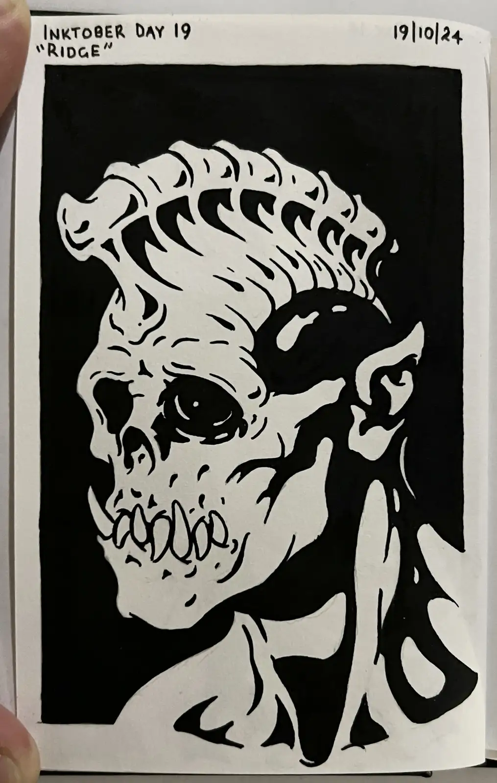 A black and white ink drawing of a demonic skull with a spiked mohawk, pointed ears, and sharp teeth, held in a sketchbook…