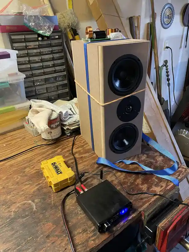 A tall, homemade speaker box with three visible drivers sits on a cluttered wooden workbench in a garage, secured with blue…
