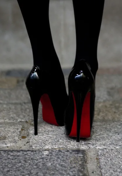 A close-up shot of a person's feet wearing glossy black stiletto heels with iconic red soles, standing on a gray stone…