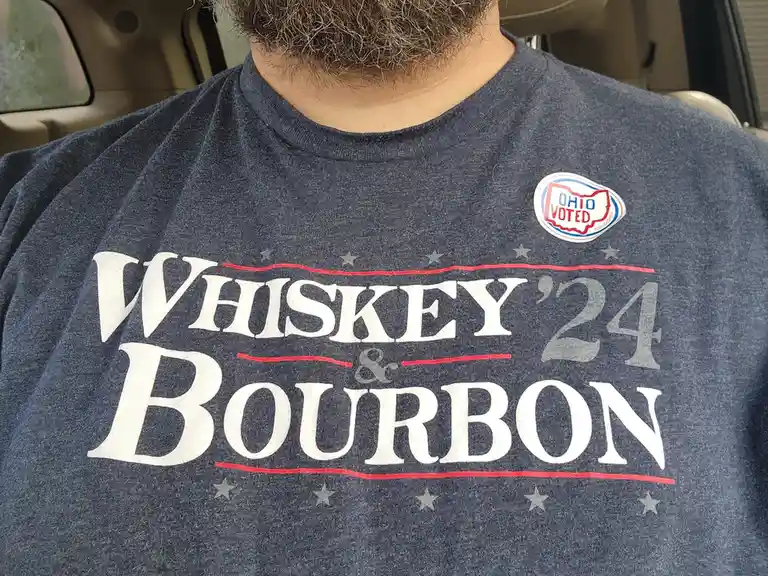 A close-up of a person's torso wearing a dark blue t-shirt with large white text reading 'WHISKEY & BOURBON '24', styled…
